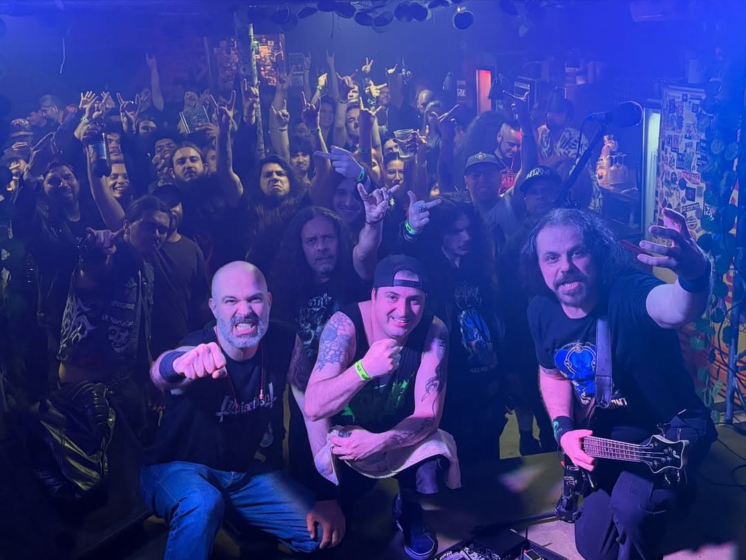 Un gruppo accovacciato davanti al piccolo palco del Caos Bar, braccia aperte, con il pubblico dietro che fa le corna sotto luci blu e viola.