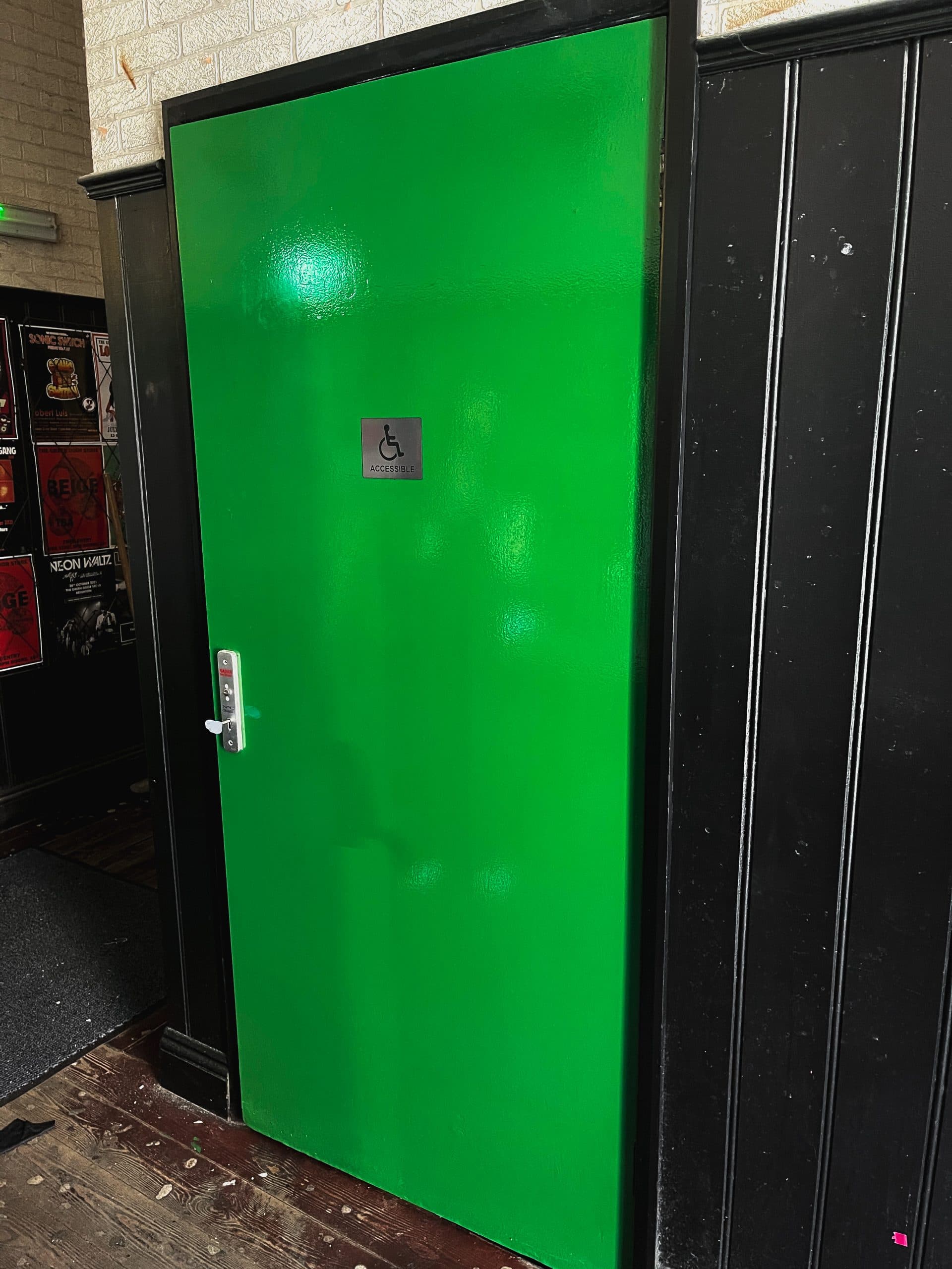 Puerta verde con señal de aseo accesible y cerradura de llave RADAR, junto a una pared llena de carteles de conciertos.