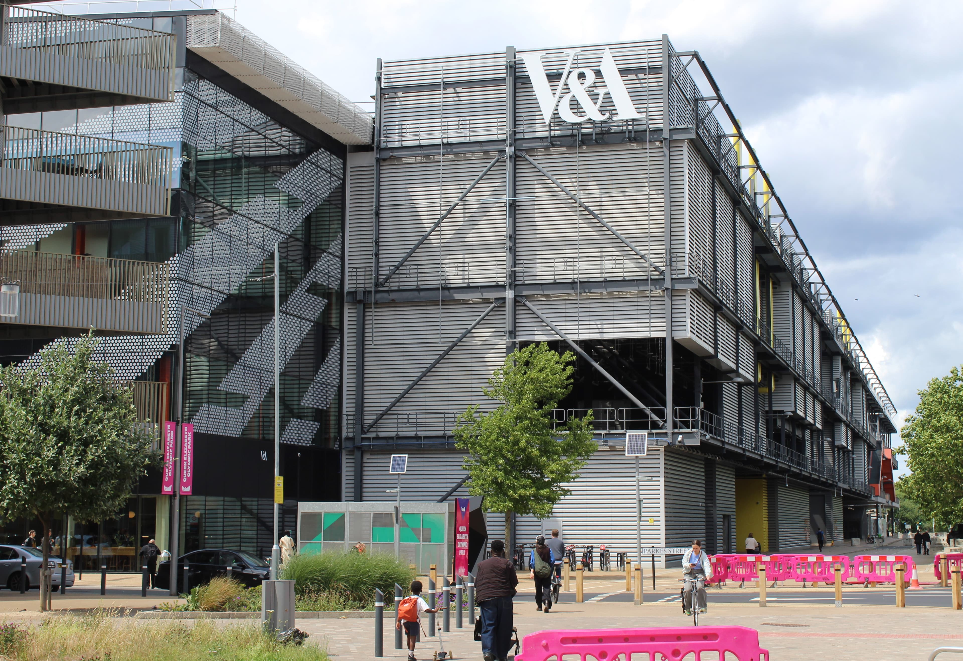 Parkes Street 上的 V&A East Storehouse 外观:一栋细长的灰色厂房,外覆金属百叶,屋顶线上有一个巨大的白色 V&A 标志。